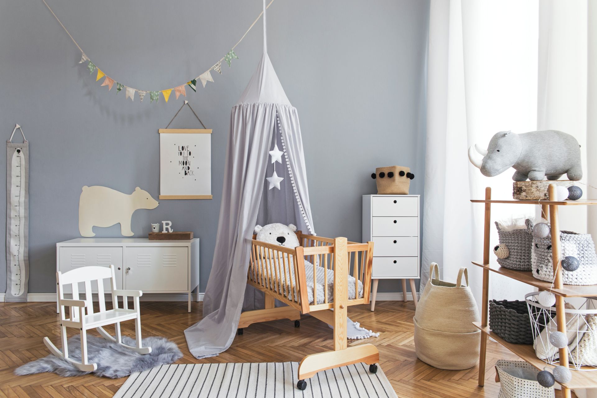 Ein liebevoll eingerichtetes Babyzimmer mit Holzkrippe, grauen Textilien, dekorativen Spielzeugen und einer weißen Schaukel – perfekte Umgebung für die Kleinsten. Baby Produkte und Einrichtungsideen für stilvolle Kinderzimmergestaltung.