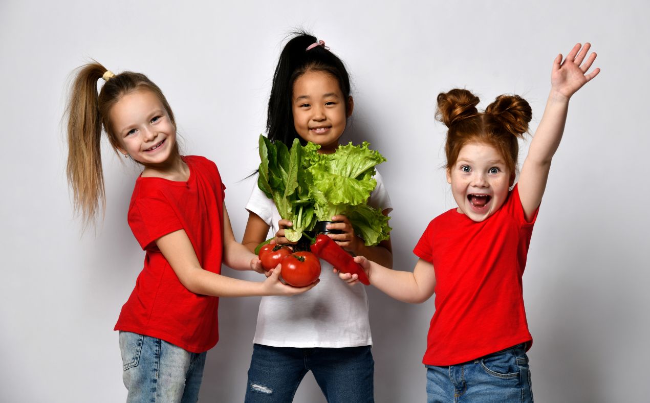 Drei lachende Kinder in roten Shirts halten frisches Gemuese wie Tomaten, Salat und Paprika – Symbol fuer gesunde Ernaehrung mit Rapunzel Naturkost