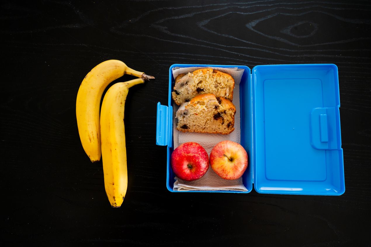 Offene Brotbox mit Bananenbrot, zwei Aepfeln und drei Bananen auf dunklem Tisch – kindgerechter Snack nach dem Vorbild von Rapunzel Naturkost