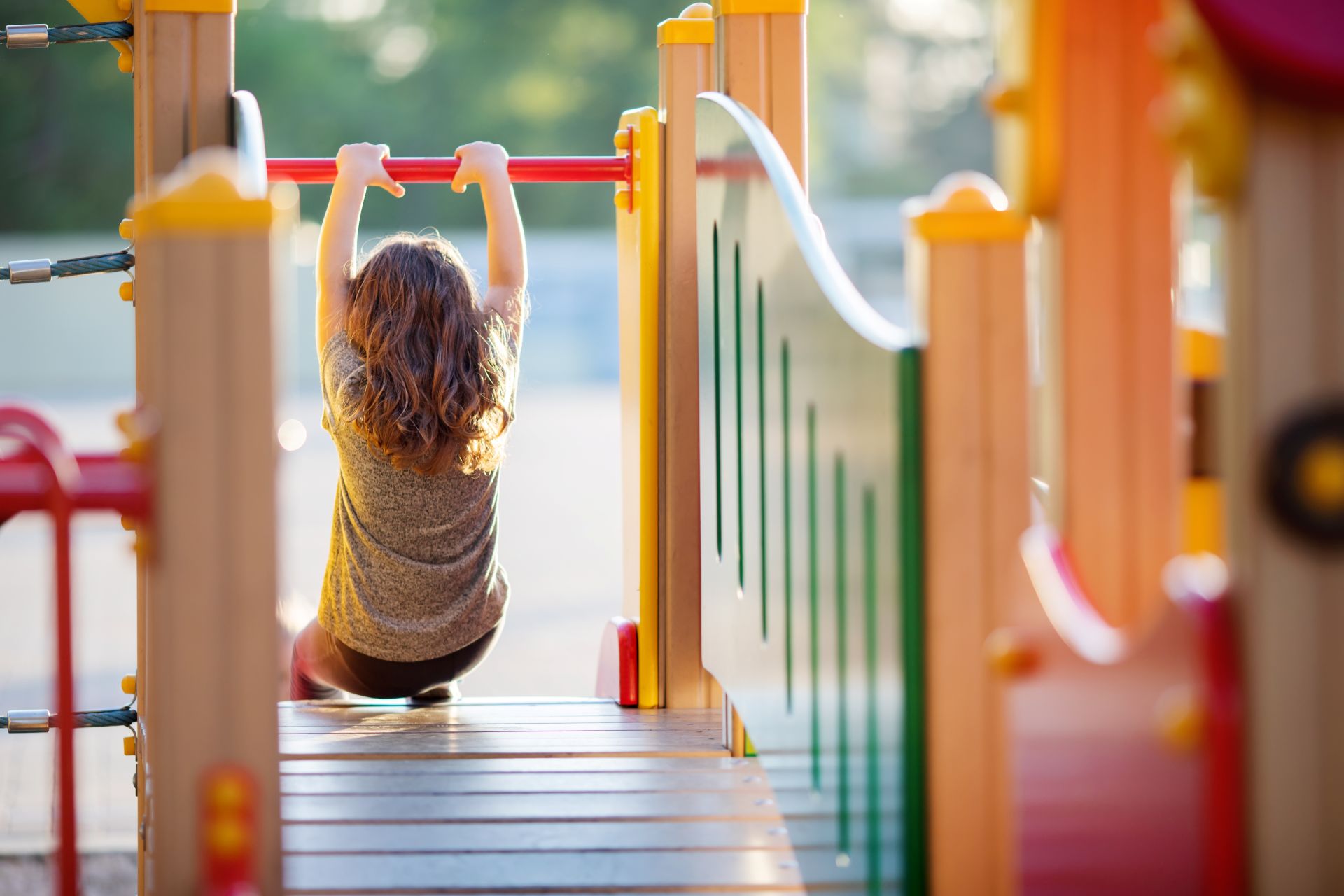 Rückansicht eines Kindes, das sich an einer roten Kletterstange auf einem bunten Spielplatz festhält.